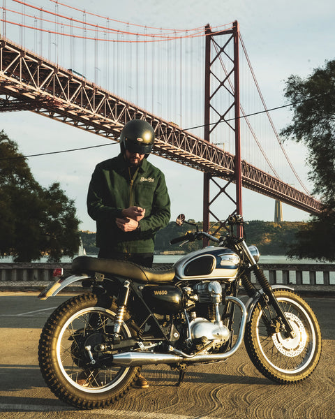 Model wearing the Bike Shed Moto Co. Twill Green logo jacket standing next to a motorcycle.