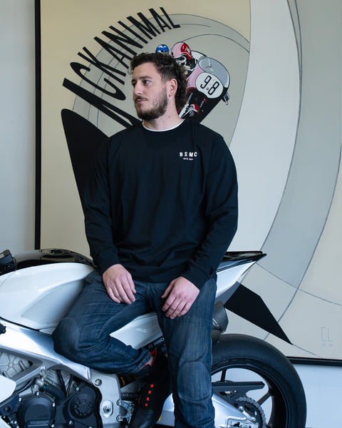 Model wearing Bike Shed Moto Co ESTD Long sleeve t-shirt in black sitting on a motorcycle in front of art work