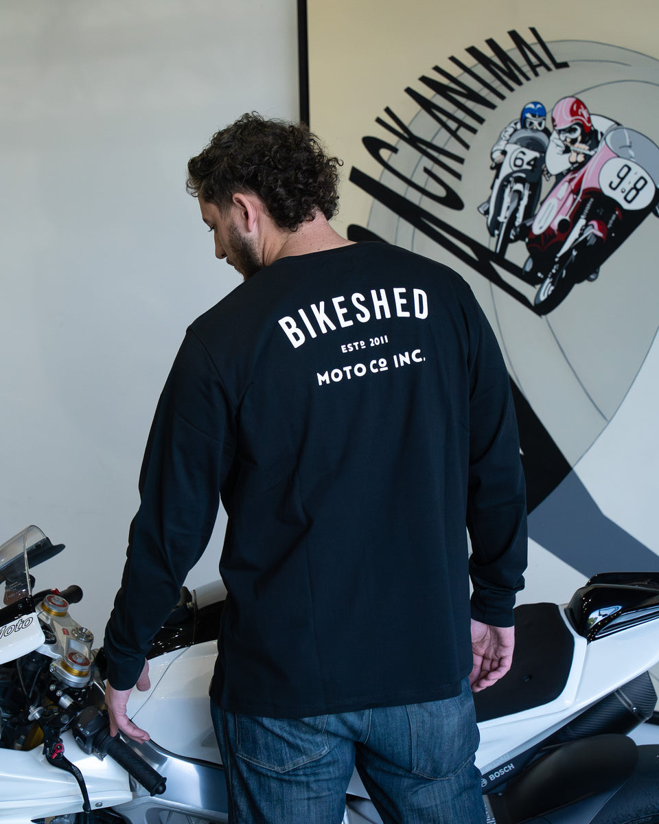 Model wearing Bike Shed Moto Co ESTD Long sleeve t-shirt in black next to a motorcycle in front of art work