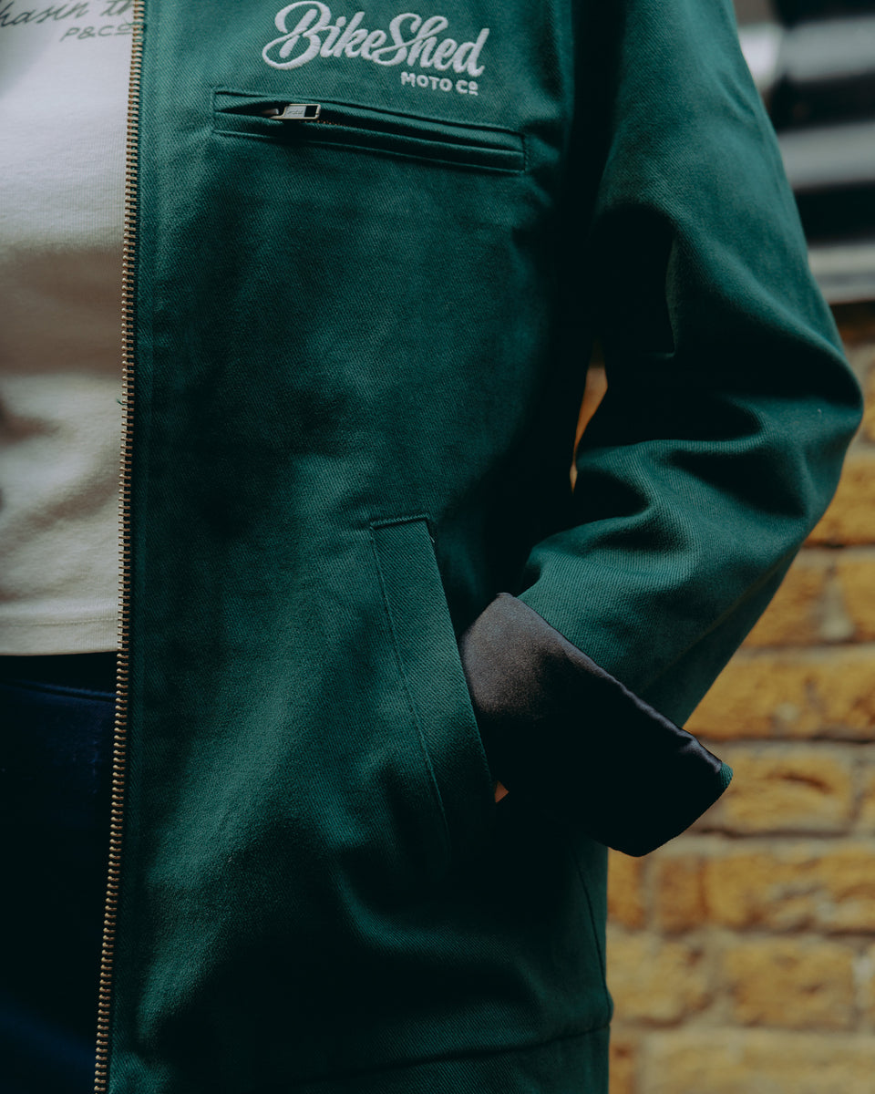 Model wearing green motorcycle jacket in front of a brick wall with graffiti.