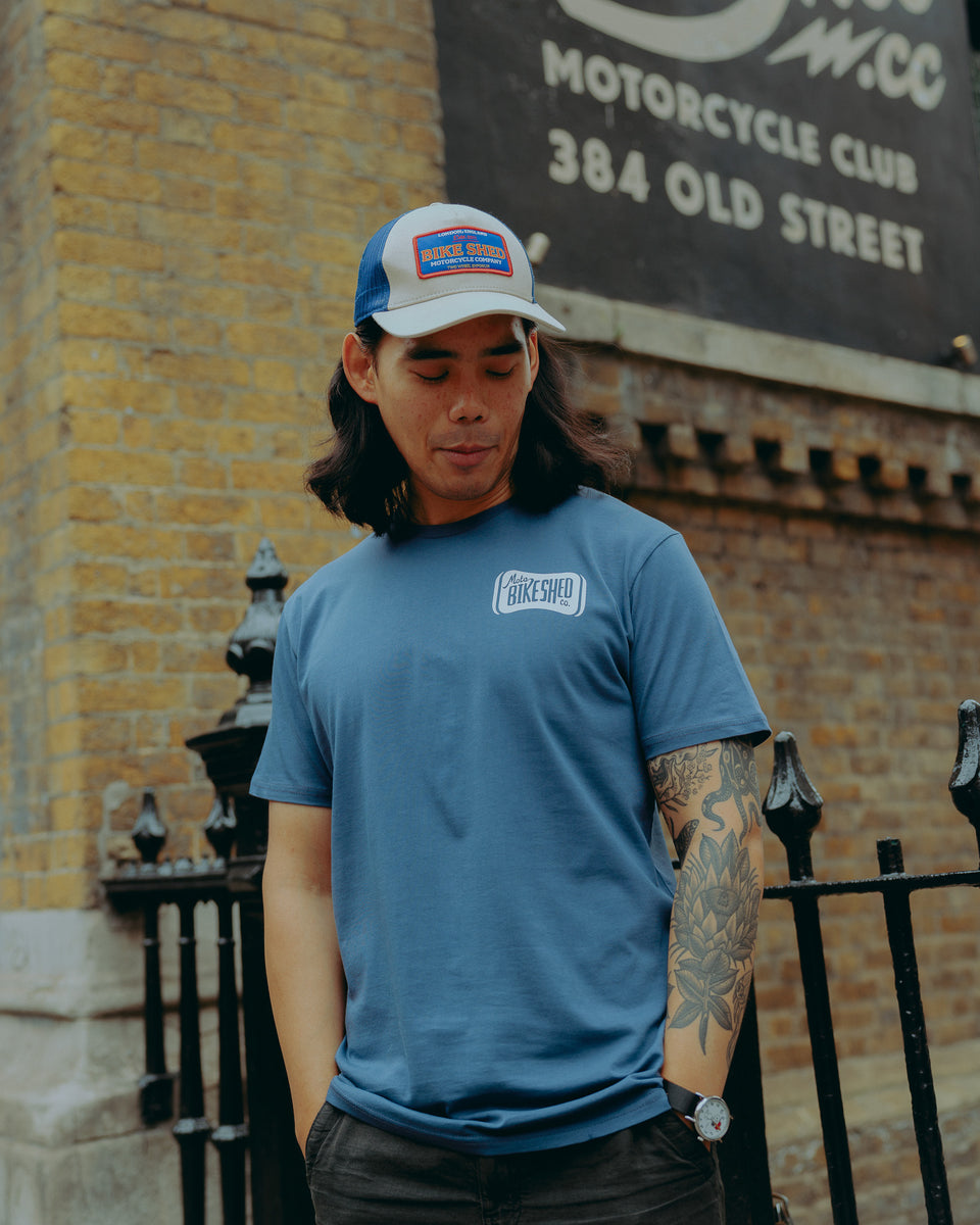 Model wearing a blue motorcycle t-shirt and cap standing in front of a brick building with a Bike Shed Moto Co sign.