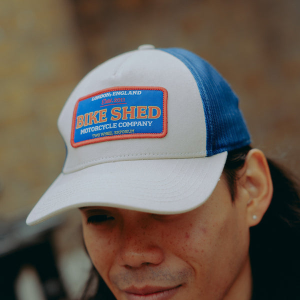 Model wearing a cream & blue motorcycle cap with 'Bike Shed Moto Co' logo against a blurred background.